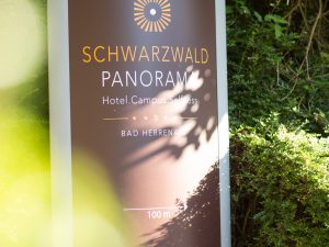 A billboard with text on it pointing towards the entrance of the hotel SCHWARZWALD PANORAMA. Text: "SCHWARZWALD PANORAMA – Hotel. Campus. Selfness. Bad Herrenalb – 100m -->"