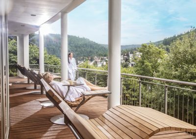 Zwei Frauen im Bademantel genießen auf einem Balkon mit Holterasse und Liegestühlen den Ausblick auf eine grüne Waldlandschaft im Hotel Schwarzwald Panorama