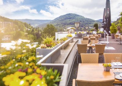 Auf einer Dachterasse stehen Tische und Stühle, im Hintergrund sieht man Wälder und die Andeutung einer Stadt, die Aussicht der Hotel Schwarzwald Panorama