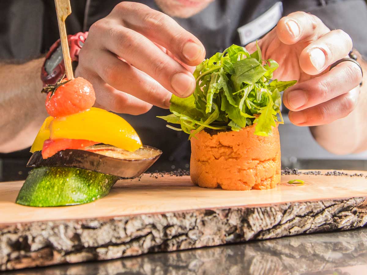Ein Koch richtet eine kunstvoll dekorierte Mahlzeit aus frischem Gemüse auf einem Holzbrett an. Eine Impression aus der Küche des Genusshotels SCHWARZWALD PANORAMA.
