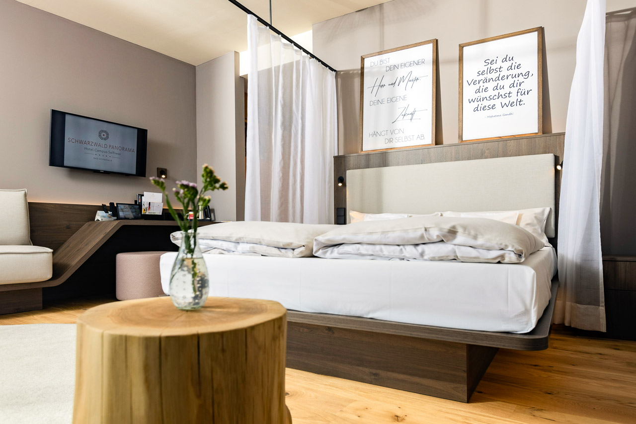 Ein Foto des CIRCULAR LIVING-Zimmers Freigeist mit Bett, Betthimmel und großen Bilderrahmen. Im Vordergrund ist ein Baumstumpf als Beistelltisch zu sehen. Im Hintergrund ein Schreibtisch mit Sitzmöglichkeit.